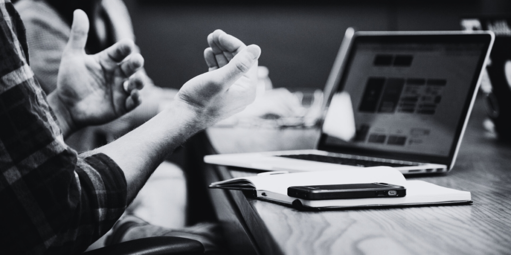 A man making hand gestures in front of a laptop, notepad and phone.