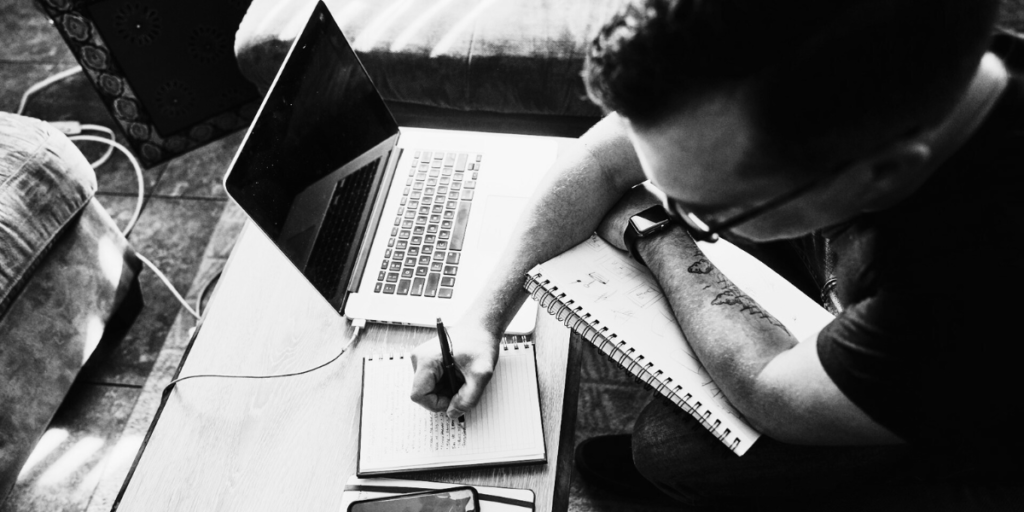 A man writing notes down on a pad with a laptop in front of him.